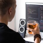 man holding clear glass mug while facing flat screen monitor